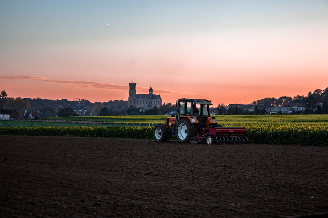 hero-homepage A red tractor plows a field under a vibrant sunset near a countryside landmark.
